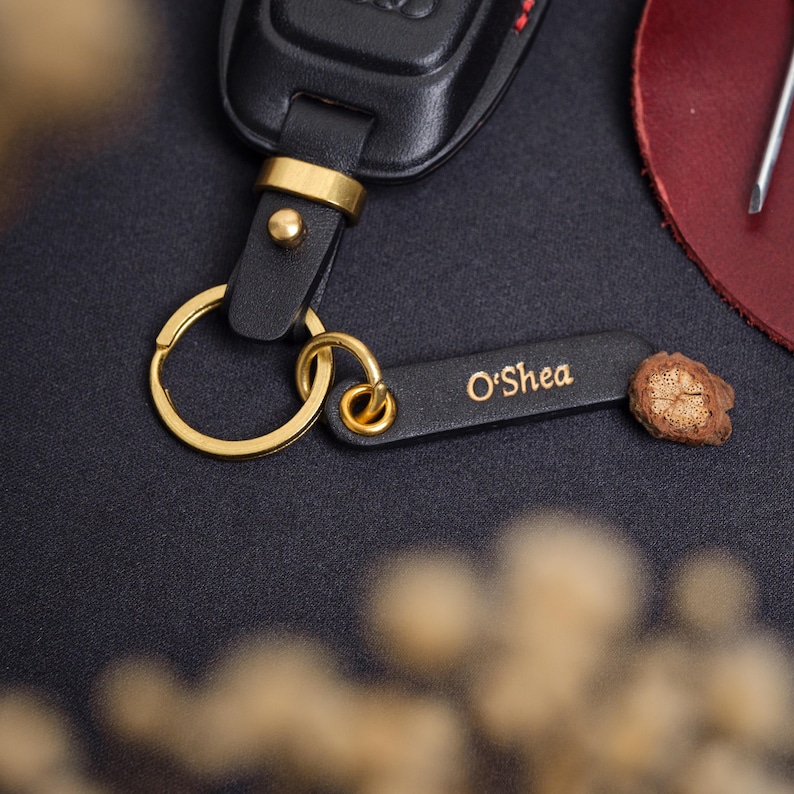 May include: A black leather key fob featuring a gold-toned metal ring and a rectangular leather tag embossed with the name O'Shea in gold lettering. The tag is attached to the ring with a gold-toned metal eyelet and a small piece of textured wood.