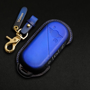 Può includere: Un portachiavi in ​​pelle blu con dettagli di cucitura neri e un logo auto argentato. Il portachiavi ha un anello portachiavi dorato con un cinturino in pelle che dice "Watson".