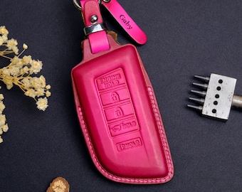 May include: A fuchsia leather key fob cover with a silver keyring and a matching pink tag that reads "Gaby". The cover has embossed buttons for "Engine Hold", lock, and unlock functions. The key fob is a vibrant accessory.