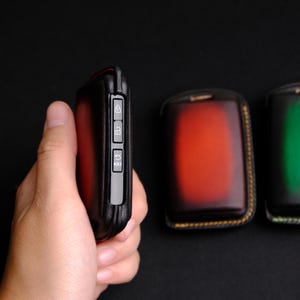 May include: Three leather key fobs with different colored leather and stitching. The key fob on the left has a black leather exterior with three buttons and the word "HOLD" printed on the button. The key fob in the middle has a red and orange leather exterior with yellow stitching. The key fob on the right has a green leather exterior with yellow stitching.