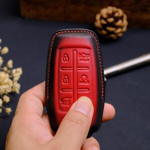 May include: A red and black leather car key fob cover featuring embossed icons for lock, unlock, hold, and trunk release functions. The cover is stitched with red thread along the edges, creating a distinct contrast against the dark leather border.