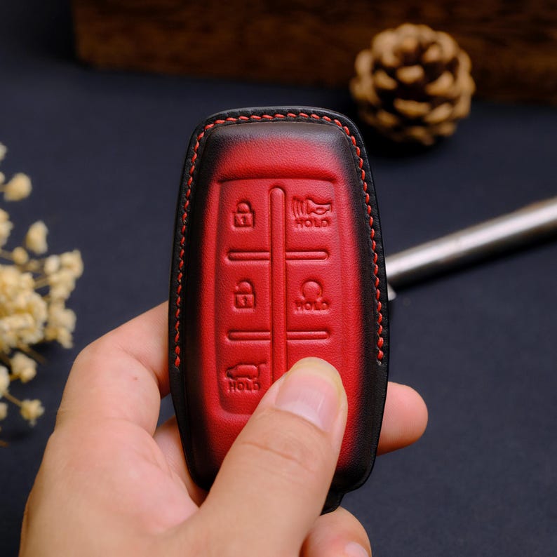 May include: A red and black leather car key fob cover featuring embossed icons for lock, unlock, hold, and trunk release functions. The cover is stitched with red thread along the edges, creating a distinct contrast against the dark leather border.