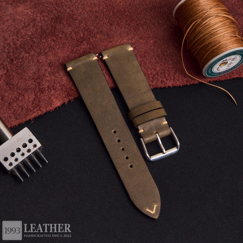 May include: Olive green leather watch strap with cream stitching and a silver buckle. The strap has a classic design with a pointed end and a series of holes for adjustment. A spool of brown thread and a leather working tool are in the background. The text "1993 LEATHER HANDCRAFTED SINCE 2022" is at the bottom.