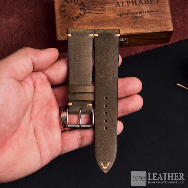 May include: Olive green leather watch strap with a silver buckle. The strap has cream-colored stitching and a V-shaped detail at the tip. The image also shows a wooden box with the word "ALPHABET" and the text "upper case & symbols (30pcs)".