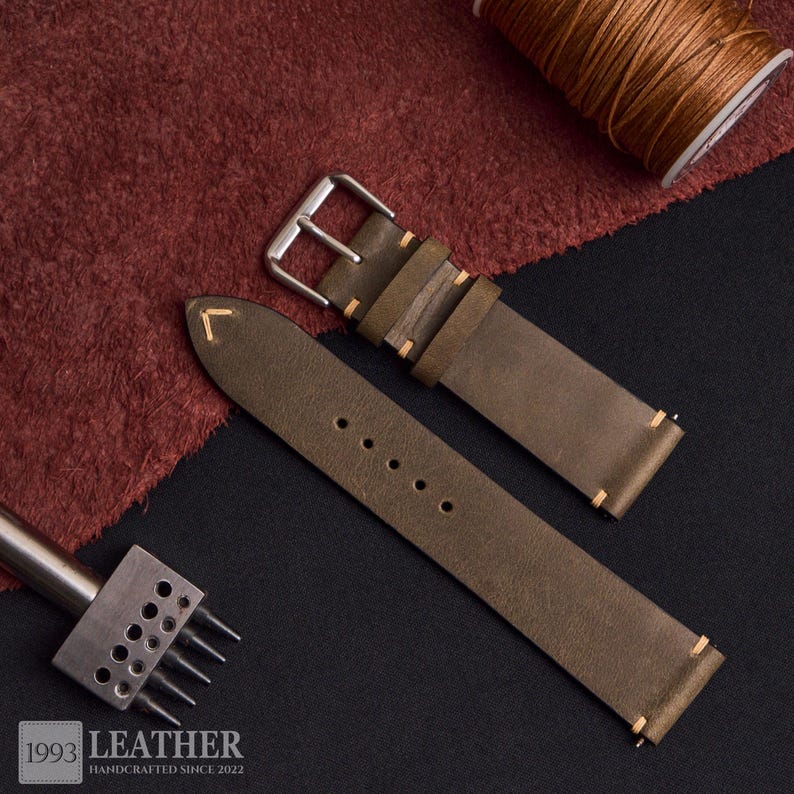 May include: Olive green leather watch strap with a silver buckle, featuring cream-colored stitching. The strap is shown with a leather crafting tool, a spool of thread, and the text "1993 LEATHER HANDCRAFTED SINCE 2022".