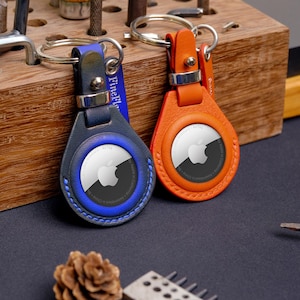 Pode incluir: Dois chaveiros AirTag de couro, um azul marinho e outro laranja, com argolas prateadas. O chaveiro azul marinho tem costuras azuis e uma borda azul ao redor do AirTag. O chaveiro laranja tem costuras laranja. Ambos têm o logotipo da Apple.