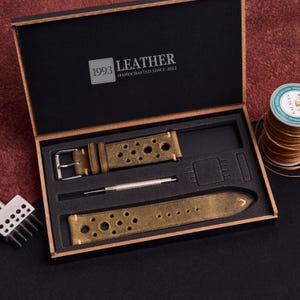 Puede incluir: Un juego de correas de reloj en una caja de madera. El juego incluye una correa de cuero verde oliva desgastado con m&uacute;ltiples agujeros, una hebilla plateada y un peque&ntilde;o destornillador. La caja tiene el texto "1993 LEATHER HANDCRAFTED SINCE 2022".