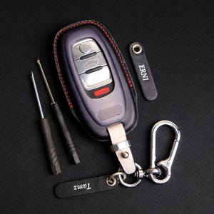 May include: A dark purple car key fob encased in a black leather cover with red stitching, featuring silver buttons and a red panic button. Included are two small screwdrivers, a silver metal carabiner, and two black leather tags labeled ERNI and Tamz.