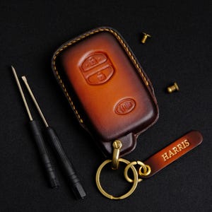 Könnte beinhalten: Ein brauner Leder-Schlüsselanhänger mit einem goldenen Schlüsselring und einem goldenen Anhänger mit der Aufschrift "HARRIS".