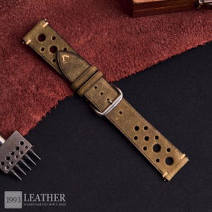 Puede incluir: Correa de reloj de cuero verde oliva desgastado con m&uacute;ltiples perforaciones circulares. La correa tiene una hebilla plateada y est&aacute; cosida con hilo de color claro. El logotipo "1993 LEATHER HANDCRAFTED SINCE 2022" es visible en la esquina inferior izquierda.