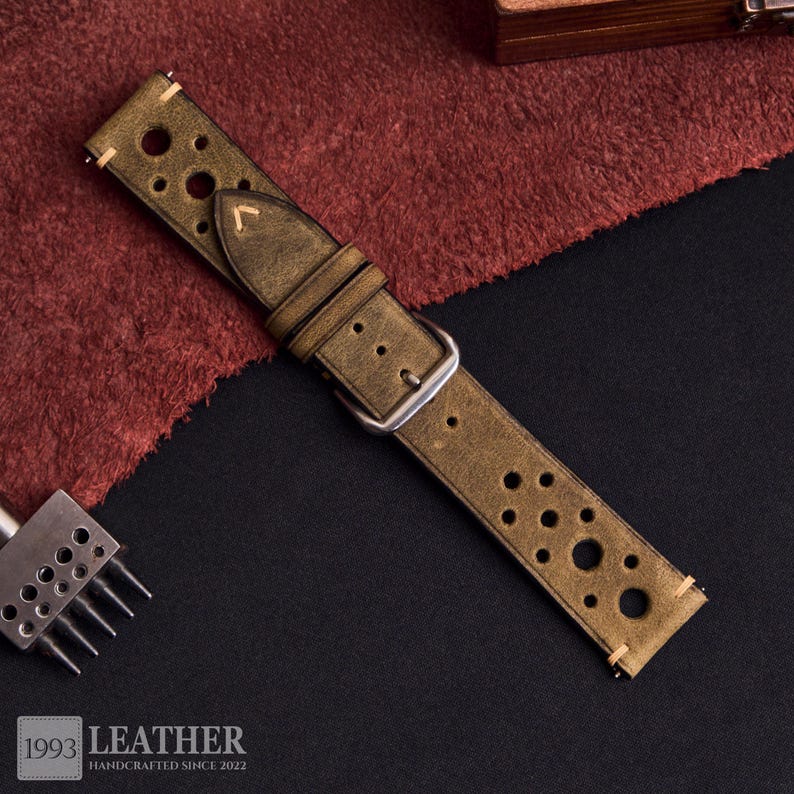 Puede incluir: Correa de reloj de cuero verde oliva desgastado con m&uacute;ltiples perforaciones circulares. La correa tiene una hebilla plateada y est&aacute; cosida con hilo de color claro. El logotipo "1993 LEATHER HANDCRAFTED SINCE 2022" es visible en la esquina inferior izquierda.