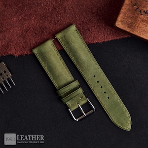 Può includere: Cinturino per orologio in pelle verde oliva con fibbia argentata. Il cinturino è realizzato in materiale scamosciato e presenta bordi cuciti. Il logo "1993 LEATHER HANDCRAFTED SINCE 2022" è visibile in basso a sinistra.