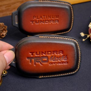 May include: Two brown leather key fob covers with black edges. One cover reads "PLATINUM TUNDRA" and the other reads "TUNDRA TRD 4x4 OFF ROAD". The covers have a stitched border and a loop for a key ring.