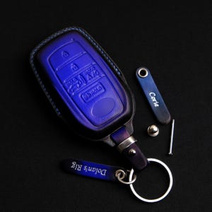 May include: A blue leather key fob with a silver key ring and a silver tag that says "Dolans' Rits". There is a smaller silver tag that says "Corle" and a silver screw.