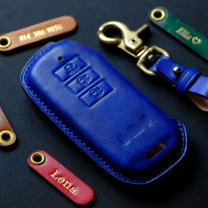 May include: A blue leather key fob with three buttons and a gold key chain. The key fob is on a black surface with other keychains with names on them.
