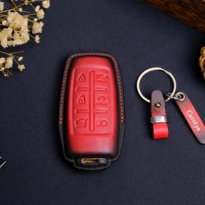 May include: A red and black gradient leather car key fob cover with embossed button icons and red stitching. Beside it is a matching leather keychain featuring a silver metal ring and a rectangular leather tag with the name Carolyn engraved.
