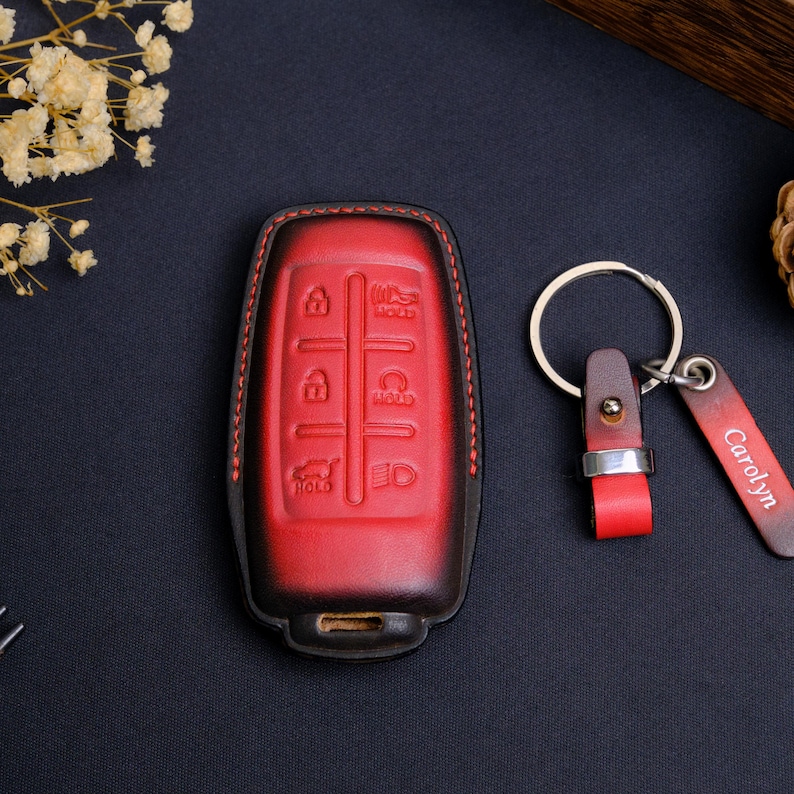 May include: A red and black gradient leather car key fob cover with embossed button icons and red stitching. Beside it is a matching leather keychain featuring a silver metal ring and a rectangular leather tag with the name Carolyn engraved.