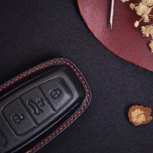 May include: A black leather car key fob cover featuring red stitching along the edges. The cover has three embossed icons representing a lock, a boot release, and an unlock function, designed to fit over a standard automotive remote key.