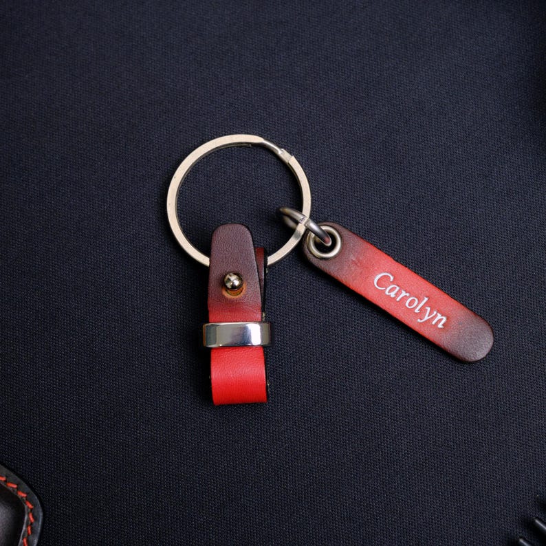 May include: A keychain featuring a circular metal ring attached to a two-tone leather strap. The strap transitions from a dark brown to a bright red, secured by a silver-toned metal rivet. A separate rectangular leather tag is engraved with Carolyn.