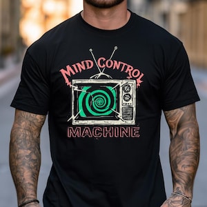 May include: A black short-sleeve t-shirt featuring a graphic of a vintage television set with an antenna. The screen displays a green spiral pattern. The text "MIND CONTROL" appears above the television and "MACHINE" below in red.