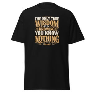 Könnte beinhalten: Ein schwarzes kurzärmeliges T-Shirt mit einem Grafikdruck in beigem und weißem Text, der lautet: THE ONLY TRUE WISDOM IS IN KNOWING YOU KNOW NOTHING - Socrates. Das Design ist mittig auf der Brust platziert und zeigt dekorative Schnörkel.