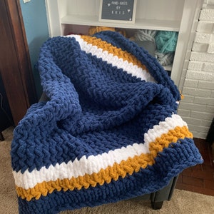 May include: A blue, white, and mustard yellow chunky knit throw blanket with a chevron pattern. The blanket is folded and draped over a chair.