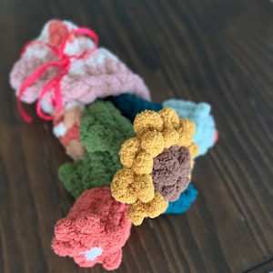 May include: A collection of colorful, handmade crochet flowers and buds. The bouquet includes a yellow sunflower, green leaves, and various buds in shades of pink, orange, and blue. A pink ribbon ties the bouquet together.