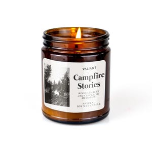 Könnte beinhalten: Ein braunes Glasgefäß mit einer Kerze und einem weißen Etikett mit der Aufschrift "Valiant Campfire Stories" und "Woody embers and crackling memories. Natural Soy Wax Candle".