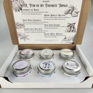 May include: A gift box set of six scented candles in silver tins. The box features text reading "A Few of My Favorite Things" and illustrations. Each candle tin has a different design on the lid, and the box is open.