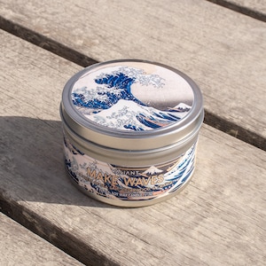 May include: A silver tin candle with a blue and white wave design on the lid. The label reads "Valiant Make Waves - Sea Salt & Cedarwood" and "11 oz Soy Wax Candle".