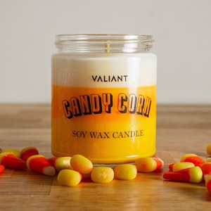 May include: A glass jar candle with a white and yellow layered wax design. The label reads "VALIANT CANDY CORN SOY WAX CANDLE". Scattered around the candle are yellow and orange candy corn pieces.