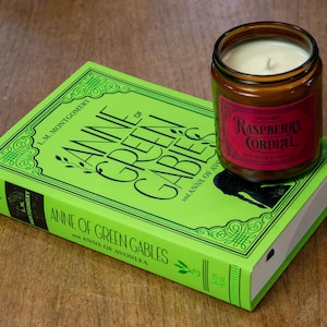 May include: A green book titled "Anne of Green Gables" and "Anne of Avonlea" by L.M. Montgomery with a brown glass candle jar on top. The candle jar has a red label that says "Raspberry Cordial".