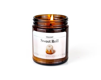 Puede incluir: Una vela en un tarro de vidrio marrón con una etiqueta blanca que dice "VALIANT Sweet Roll" y presenta una pequeña ilustración de un rollo dulce. La vela está encendida y la llama es visible.