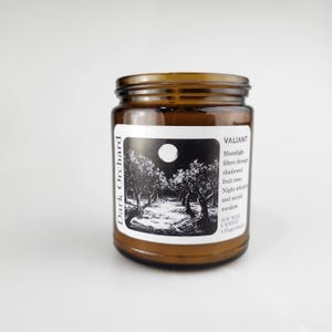 Könnte beinhalten: Ein braunes Glas-Kerzenglas mit einem weißen Etikett, das eine Schwarz-Weiß-Illustration von Bäumen unter einem Vollmond zeigt. Das Etikett trägt die Aufschrift "Dark Orchard" und "VALIANT". Die Kerze ist eine Sojawachskerze.