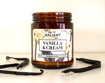 Może przedstawiać: Świeca w brązowym szklanym słoiku z białą etykietą, na której widnieje napis "VALIANT VANILLA & CREAM NO. 23 HANDMADE IN UTAH 8 FL. OZ.". Etykieta przedstawia botaniczny rysunek waniliowych strąków i kwiatów.