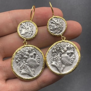 May include: A pair of silver and gold dangle earrings featuring two round coin-shaped charms with detailed engravings of ancient Roman emperors' profiles.