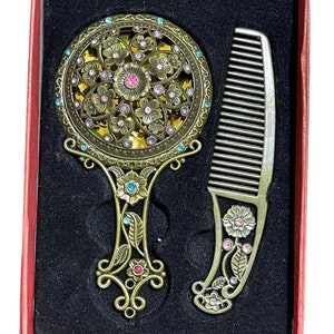 May include: A gold-toned vanity set with a floral design. The set includes a hand mirror with a round, ornate frame and a comb with a decorative handle. Both pieces are adorned with rhinestones.