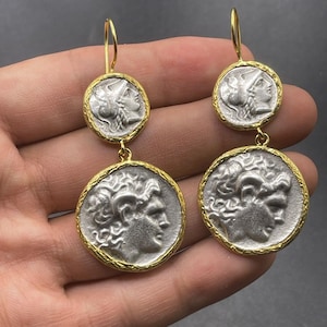 May include: A pair of silver-toned earrings with gold-colored accents. Each earring features two circular pendants with embossed classical figures. The figures are set within a gold-colored frame, and the earrings have a gold-colored hook.