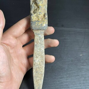 May include: A long, thin, silver-coloured metal object with a small, gold-coloured figure at the top. The figure is seated and appears to be wearing a crown. The object is decorated with intricate carvings.