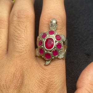 Może przedstawiać: Srebrny pierścionek w kształcie żółwia z centralnym owalnym rubinem otoczonym mniejszymi, okrągłymi rubinami. Głowa i nogi żółwia są szczegółowe. Pierścionek jest noszony na palcu.