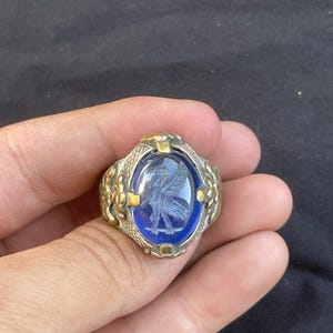 Peut inclure: Une bague en argent avec une pierre ovale bleue sertie dans un cadre en métal doré. La pierre est gravée d'une figure ailée.