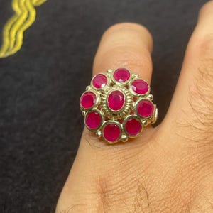 Może przedstawiać: Srebrny pierścionek z kwiatowym wzorem, z centralnym owalnym rubinem otoczonym przez dziesięć okrągłych rubinów. Pierścionek jest prezentowany na palcu, podkreślając jasnoczerwone kamienie szlachetne.