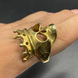 May include: A gold-toned ring featuring a skull design with a crown on top. The ring is worn on a finger.