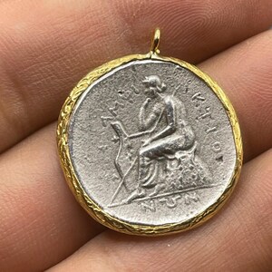 May include: A silver coin pendant with a gold rim. The coin features a seated figure with a crown and a staff. The coin is inscribed with the text "AM", "ANT", and "KAI".