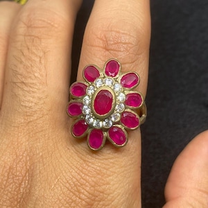 Może przedstawiać: Złoty pierścionek z kwiatowym wzorem. Środek pierścionka to duży owalny rubin, otoczony małymi białymi kamieniami szlachetnymi. Rubiny w kształcie płatków otaczają środek, tworząc kwiatowy wygląd.