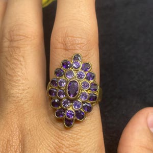 Może przedstawiać: Złoty pierścionek z owalnym, ciemnofioletowym kamieniem centralnym otoczonym mniejszymi, okrągłymi, fioletowymi kamieniami szlachetnymi. Projekt pierścionka to skupisko kamieni, tworzące wzór kwiatowy lub gwiazdowy. Pierścionek jest noszony na palcu.