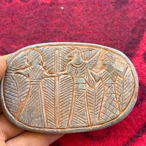Puede incluir: Un óvalo de piedra gris con un diseño tallado de cuatro figuras en relieve. Las figuras llevan túnicas y tocados. El diseño es intrincado y detallado.