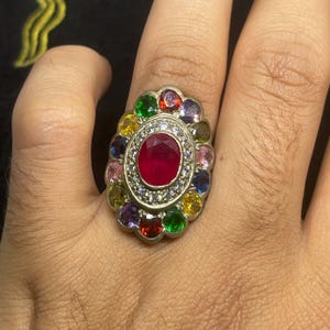 Może przedstawiać: Srebrny pierścionek z dużym owalnym rubinem w środku, otoczonym aureolą małych białych kamieni. Pierścionek jest ozdobiony szeregiem mniejszych kamieni szlachetnych w kolorach takich jak czerwony, zielony, żółty, niebieski i fioletowy.