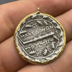 May include: A silver coin pendant with a gold rim. The coin features an inscription in Greek, "ΜΑΚΕΔΟΝΩΝ" and "ΠΡΟΣΘΗ", surrounded by a wreath of leaves.