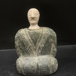 Puede incluir: Una pequeña escultura de piedra verde de una figura sentada con una cabeza de piedra blanca separada. La figura lleva una prenda con un patrón tallado.